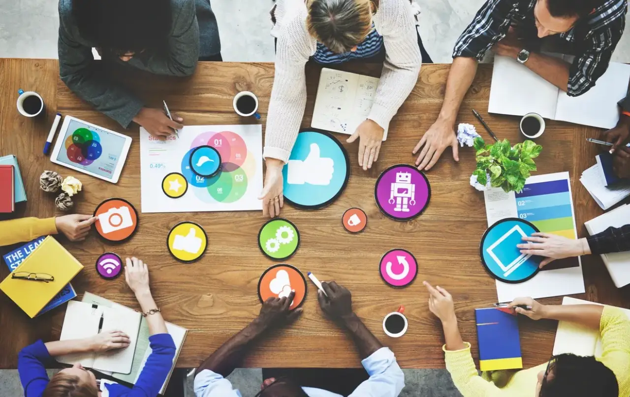 A diverse group collaborates around a wooden table, discussing ideas with colorful icons, notebooks, and drinks
